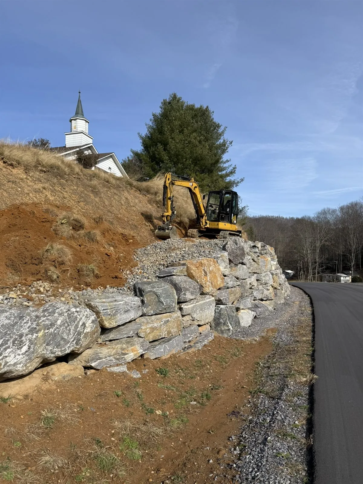 Boulder retaining wall built along a paved road below a hilltop church, excavator on top, Western NC