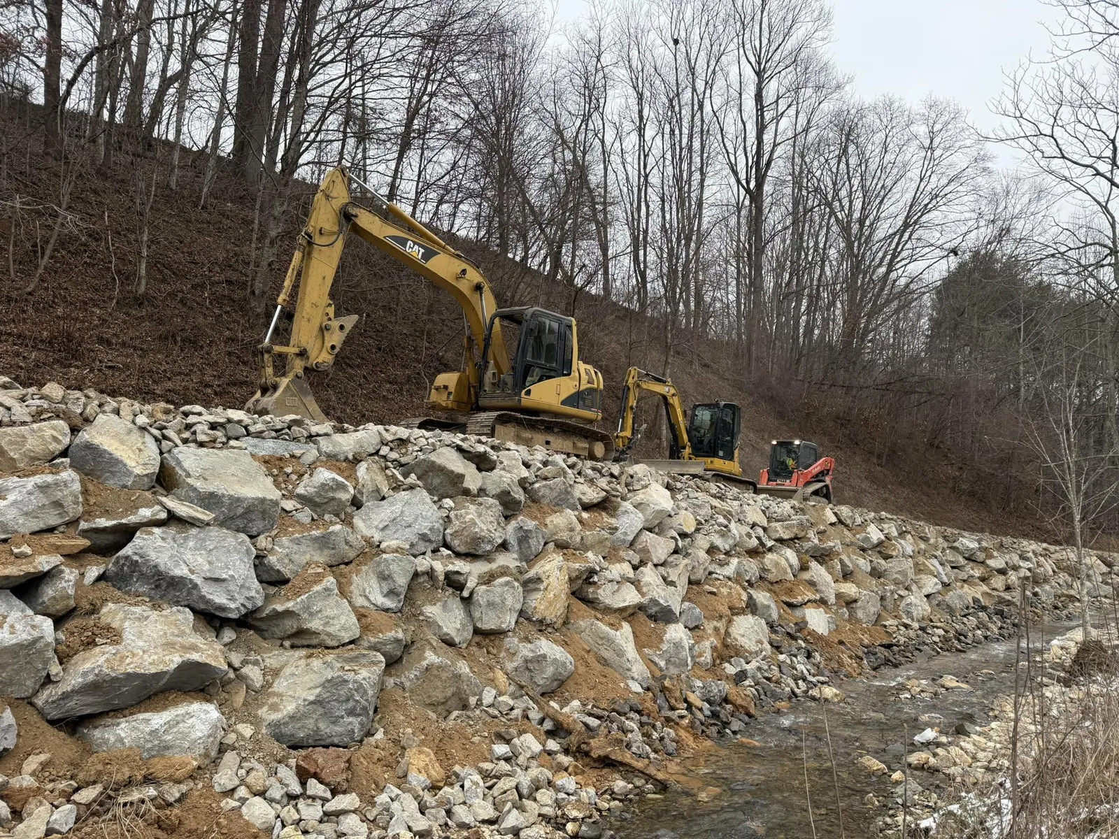 Excavator and skid steer building a boulder and riprap retaining wall along a creek