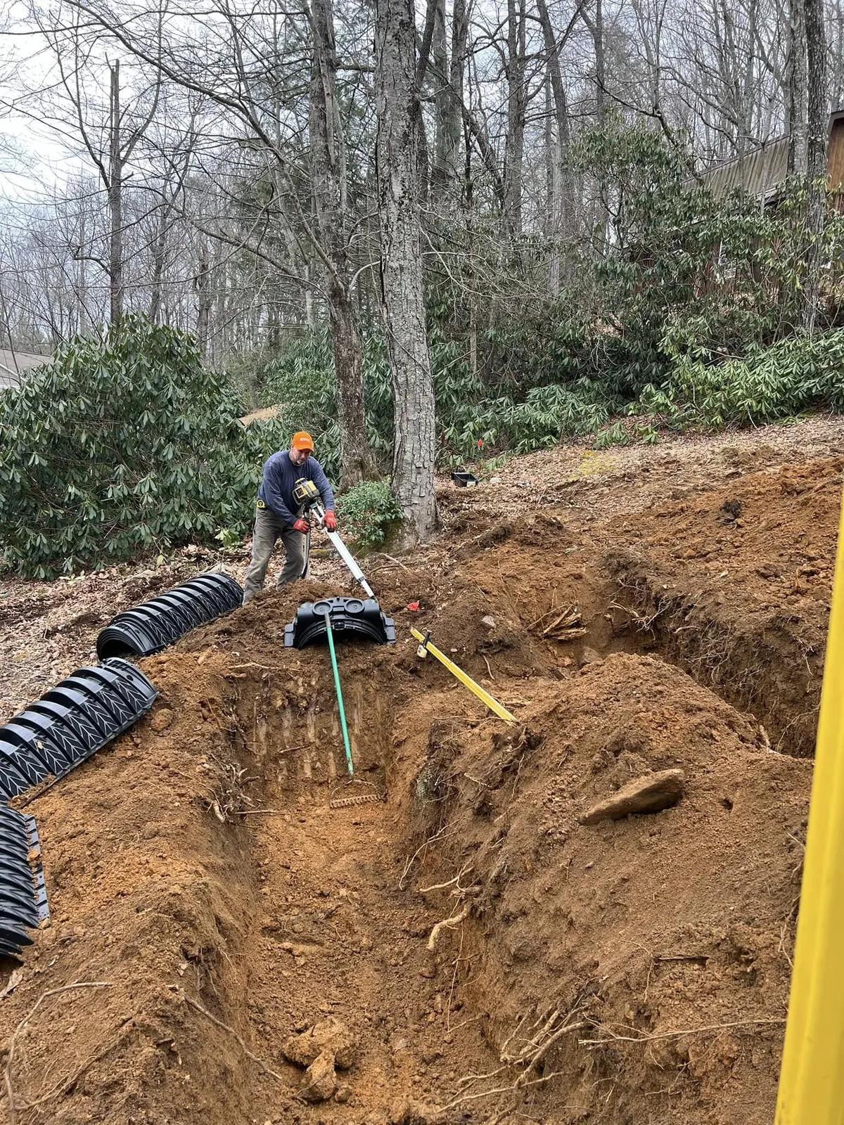 Worker laying corrugated drainage pipe in a trench on a wooded WNC slope